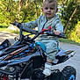 child, toddler, atv, toy_vehicle, outdoor, trees, greenery, sunlight, road, smiling, face, clothing, shoes, handlebars, wheels, nature, adventure, play, vehicle, young_child