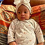 baby, infant, headband, bow, onesie, patterned_clothing, blanket, colorful, indoor, cute, child, portrait, relaxed, young, person, small_hands, face, expression, seated, soft_lighting