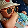 baby, child, sunglasses, hat, strawberry_patch, thumb_sucking, car_seat, seatbelt, plush_toy, floral_clothing, infant, cute, orange_glasses, blue_toy, pacifier, relaxed, indoor, portrait, casual, comfortable