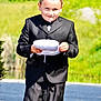 James is registered to the contest to win money with this photo: child, boy, suit, black_suit, white_pillow, outdoor, greenery, smiling, portrait, formal_wear, young_boy, person, nature, sunlight, grass, pathway, happy, cute, short_hair, daytime