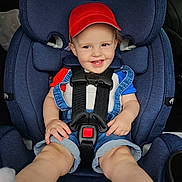 Colton is registered to the contest to win money with this photo: baby, blue_clothing, car, car_seat, casual, child, cute, denim_overalls, happy, infant, interior, person, portrait, red_cap, safety_harness, seat, seatbelt, smiling, toddler, transportation