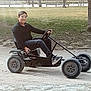 Sidi Mikhail participe au concours pour gagner de l'argent avec cette photo : child, boy, go_kart, steering_wheel, outdoor, park, grass, trees, bench, casual_clothing, black_shirt, sneakers, smiling, daylight, recreational_vehicle, play, youth, person, fun, leisure