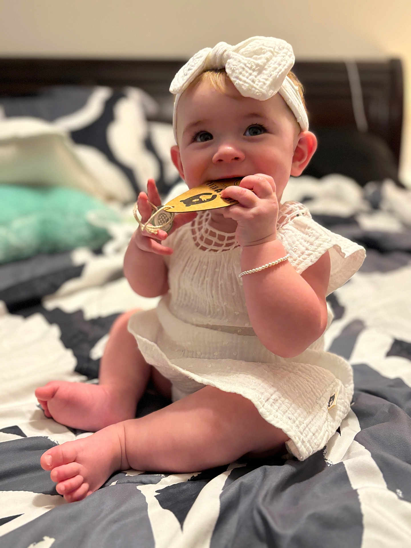 Lydia is registered to the contest to win money with this photo: baby, child, infant, white_dress, headband, bow, bed, bedding, pillow, tag, chewing, cute, smiling, sitting, skin, hand, foot, bracelet, indoors, soft_light