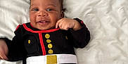 Jasiah is registered to the contest to win money with this photo: baby, infant, smiling, black_clothing, gold_buttons, white_belt, lying_down, bed, pillow, happy, cute, child, young, face, person, indoors, soft, blanket, blue_pants, playful