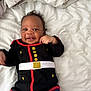 baby, infant, smiling, black_clothing, gold_buttons, white_belt, lying_down, bed, pillow, happy, cute, child, young, face, person, indoors, soft, blanket, blue_pants, playful