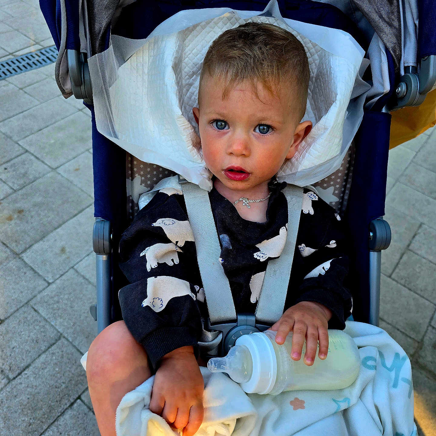 Nikita is registered to the contest to win money with this photo: baby, baby_bottle, blanket, blue_eyes, child, clothing, curly_hair, expression, face, foot, hand, necklace, outdoor, pavement, person, seat, straps, stroller, sunlight, toddler