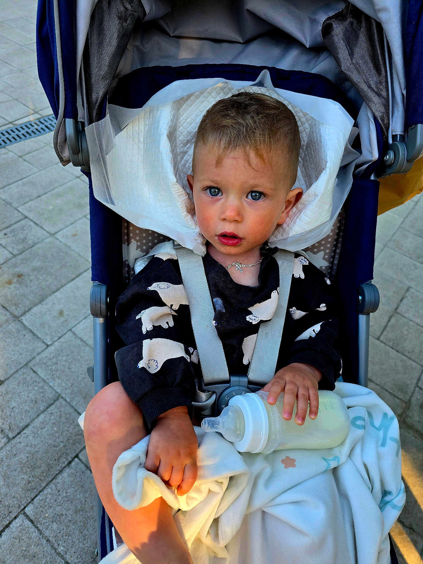 Nikita is registered to the contest to win money with this photo: baby, baby_bottle, blanket, blue_eyes, child, clothing, curly_hair, expression, face, foot, hand, necklace, outdoor, pavement, person, seat, straps, stroller, sunlight, toddler