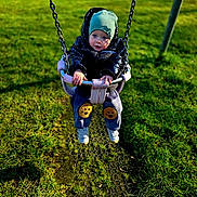 Nikita is registered to the contest to win money with this photo: child, toddler, swing, playground, jacket, beanie, grass, outdoor, park, baby, hands, face, footwear, puffy_jacket, pants, cookie_patch, seat, chain, fun, cute