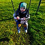 child, toddler, swing, playground, jacket, beanie, grass, outdoor, park, baby, hands, face, footwear, puffy_jacket, pants, cookie_patch, seat, chain, fun, cute