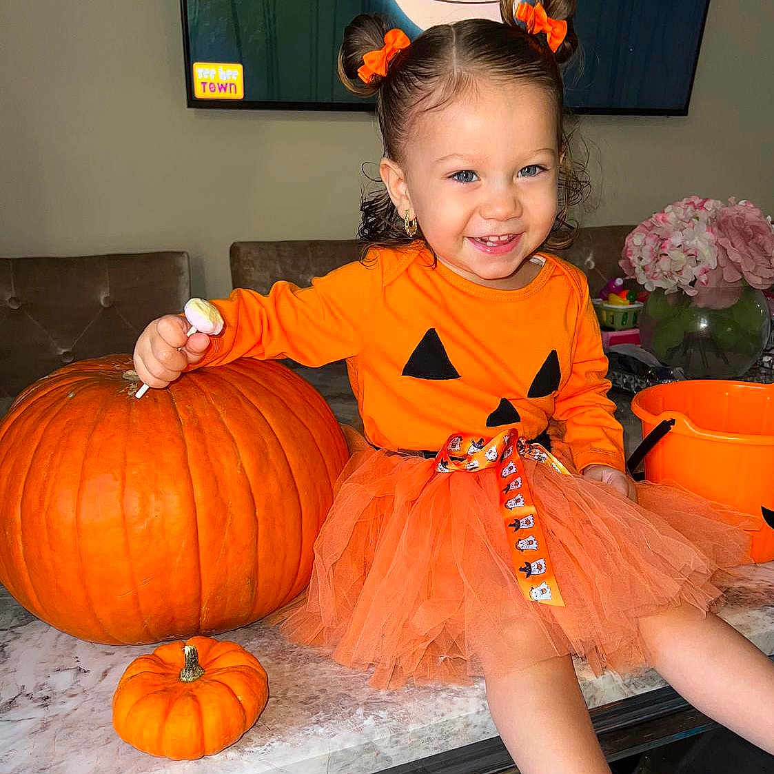Monroe Blu is registered to the contest to win money with this photo: bucket, child, cute, decoration, dress, festive, flower_vase, flowers, hair_bows, happy, holiday, indoor, lollipop, marble_table, orange, pumpkin, smile, table, toddler, tutu