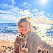 Céleste a rejoint le concours — aidez-le/la à gagner de superbes lots ! child, smiling, beach, ocean, sunlight, sky, clouds, water, jacket, cozy, happy, outdoor, nature, seashore, sand, waves, portrait, sun, holiday, warm_clothing