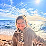 child, smiling, beach, ocean, sunlight, sky, clouds, water, jacket, cozy, happy, outdoor, nature, seashore, sand, waves, portrait, sun, holiday, warm_clothing