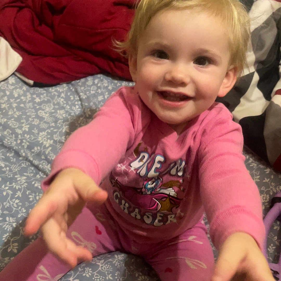 Harlenn joined the competition — help win amazing prizes! bed, blanket, blond_hair, child, cozy, cute, face, foot, happy, home, indoor, person, pink_pajamas, plush_toy, reaching, sitting, sleepwear, smiling, toddler, young_child