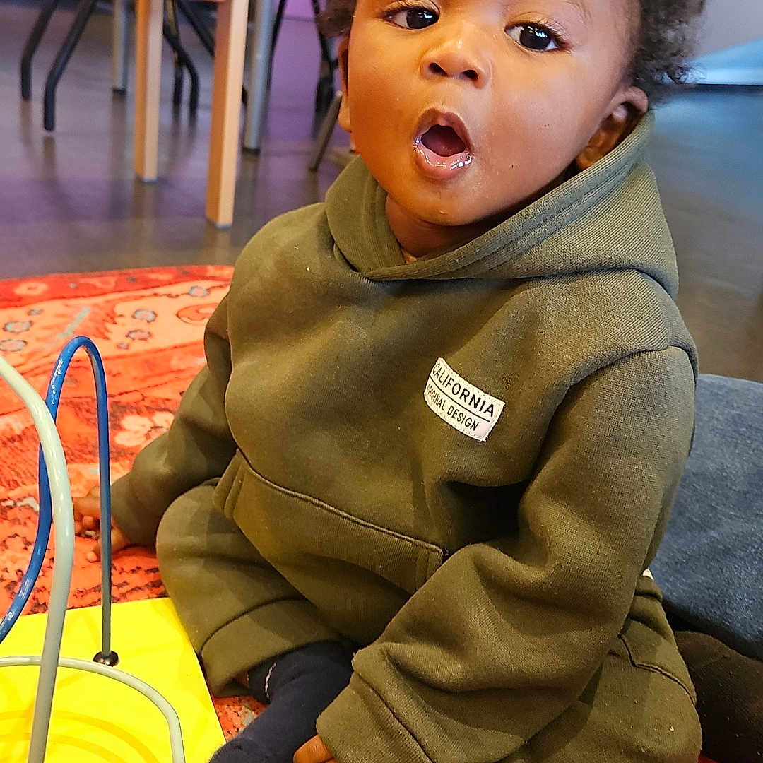 Zayonn participe au concours pour gagner de l'argent avec cette photo : background, carpet, casual_clothing, child, colorful, curious, curly_hair, expression, floor, furniture, home, hoodie, indoor, person, play_area, seated, socks, toddler, toy, young_child