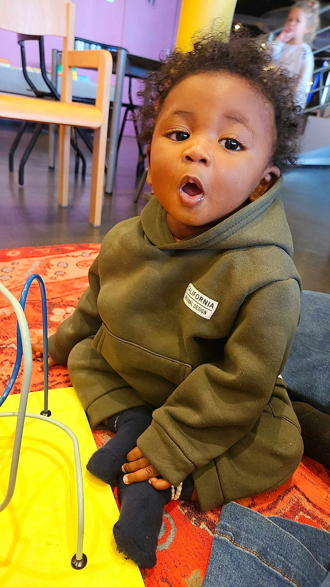 Zayonn participe au concours pour gagner de l'argent avec cette photo : background, carpet, casual_clothing, child, colorful, curious, curly_hair, expression, floor, furniture, home, hoodie, indoor, person, play_area, seated, socks, toddler, toy, young_child