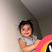 Nashly joined the competition — help win amazing prizes! toddler, child, smiling, happy, girl, pigtails, hair_bows, gray_shirt, patterned_pants, indoor, wall, portrait, cute, baby, person, skin, face, holding, colorful_object, playful