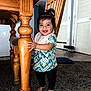Nashly is registered to the contest to win money with this photo: toddler, child, indoor, carpet, wooden_table, wooden_chair, table_leg, smiling, barefoot, bib, short_sleeve_shirt, black_pants, hair_tied, happy, standing, home, floor, door, furniture, person