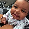 Amara is registered to the contest to win money with this photo: baby, infant, child, smiling, happy, portrait, close_up, face, eyes, indoors, bed, bedding, patterned_background, floral_clothing, onesie, sitting, leaning, adult_arm, candid, photography