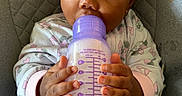 Amara is registered to the contest to win money with this photo: baby, infant, child, bottle, feeding, milk, sitting, chair, portrait, close_up, face, eyes, hands, onesie, floral_clothing, purple, indoor, lounge_chair, cute, photography