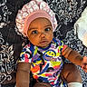Amara is registered to the contest to win money with this photo: baby, infant, child, portrait, lying_down, sitting, wide_eyed, serious_expression, bonnet, pink_hat, colorful_clothing, onesie, socks, blanket, bed, indoor, close_up, dark_skin, patterned_background, cute