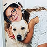 child, dog, helmet, glasses, hug, indoor, white_background, smile, pet, friendship, happy, curly_hair, casual_clothing, floor_tiles, closeup, cute, affection, young_girl, canine, domestic_animal