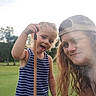 child, person, outdoor, grass, stick, cap, striped_clothing, smile, tongue_out, fun, young, casual, nature, daytime, two_people, portrait, happy, playful, summer, field