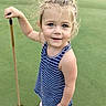 River Grace is registered to the contest to win money with this photo: child, girl, blue_striped_dress, golf, putter, golf_hole, green_grass, outdoor, standing, smiling, blonde_hair, casual_clothing, sport, recreation, young_child, summer, playful, portrait, daytime, nature