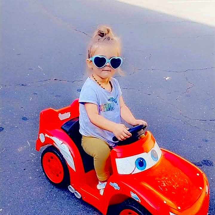 River Grace is registered to the contest to win money with this photo: child, toddler, sunglasses, toy_car, red, parking_lot, outdoor, asphalt, casual_clothing, blond_hair, person, vehicle, daytime, fun, play, cute, summer, small, sitting, happy