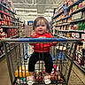 child, toddler, baby, grocery_store, shopping_cart, supermarket, sitting, smiling, portrait, red_shirt, black_pants, floral_shoes, food, shelves, condiments, barbecue_sauce, yogurt, shopping, indoor, family_outing