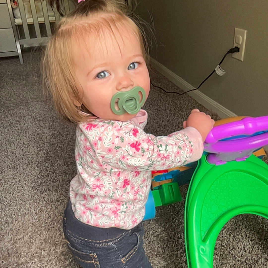 Ellie is registered to the contest to win money with this photo: toddler, child, baby, portrait, pacifier, blonde_hair, ponytail, blue_eyes, smiling, standing, indoor, home, nursery, crib, carpet, toy, walker, colorful, floral_shirt, jeans