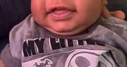 Christian joined the competition — help win amazing prizes! baby, smiling, infant, tshirt, indoor, child, portrait, happy, face, person, dark_background, phone_screen, text_overlay, hand, closeup, cute, young_child, casual_clothing, sitting, expression