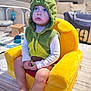 toddler, child, plush_chair, green_hoodie, dinosaur_costume, blue_eyes, indoor, carpet, toy, furniture, living_room, soft_lighting, cute, sitting, barefoot, expression, young_child, casual, portrait, cozy