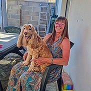 Pralinette participe au concours pour gagner de l'argent avec cette photo : woman, dog, outdoor, plastic_chair, smiling, glasses, long_hair, patterned_dress, happy, pet, sunlight, brick_wall, ladder, table, pavement, animal, person, sitting, tongue_out, garden