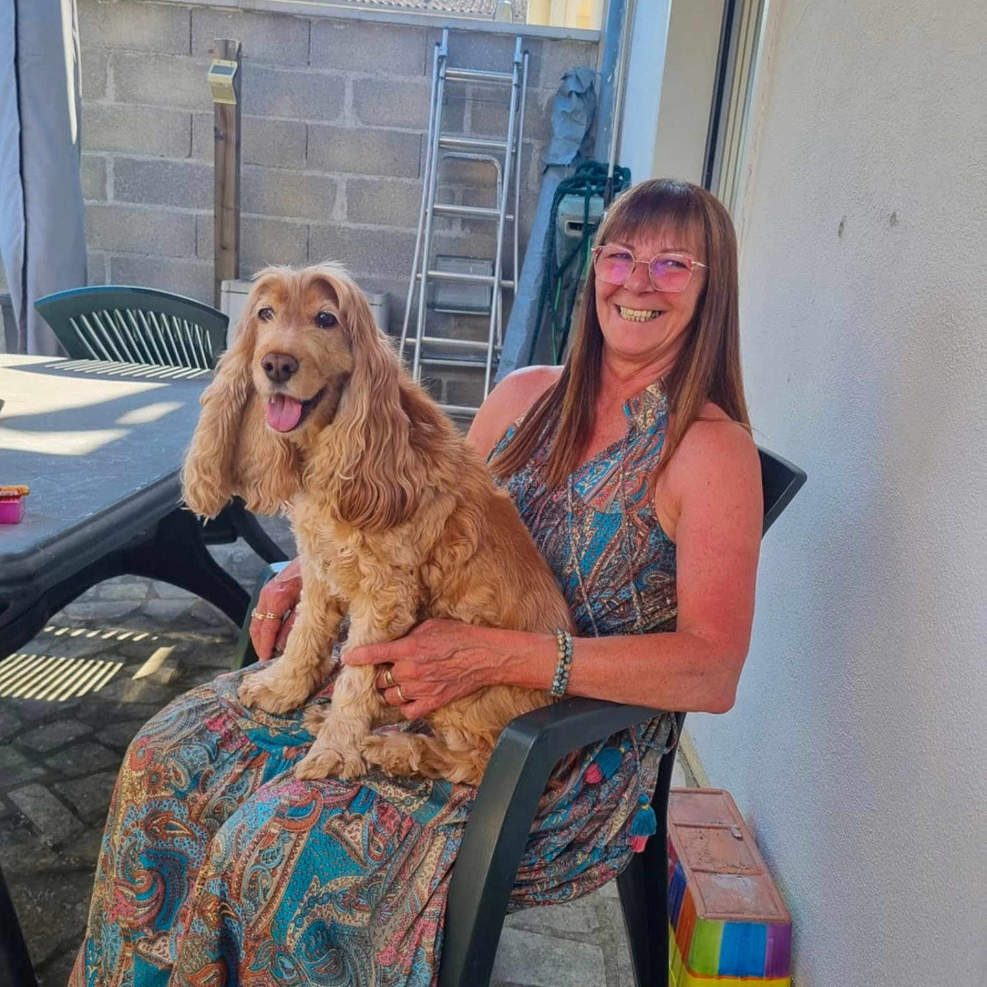 Pralinette participe au concours pour gagner de l'argent avec cette photo : animal, brick_wall, dog, garden, glasses, happy, ladder, long_hair, outdoor, patterned_dress, pavement, person, pet, plastic_chair, sitting, smiling, sunlight, table, tongue_out, woman