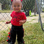 Emoni is registered to the contest to win money with this photo: toddler, child, smile, red_bow, red_shirt, black_pants, ribbons, hair_beads, outdoor, grass, chain_link_fence, sunny_day, sidewalk, trees, happy, person, portrait, fashion, cute, playful