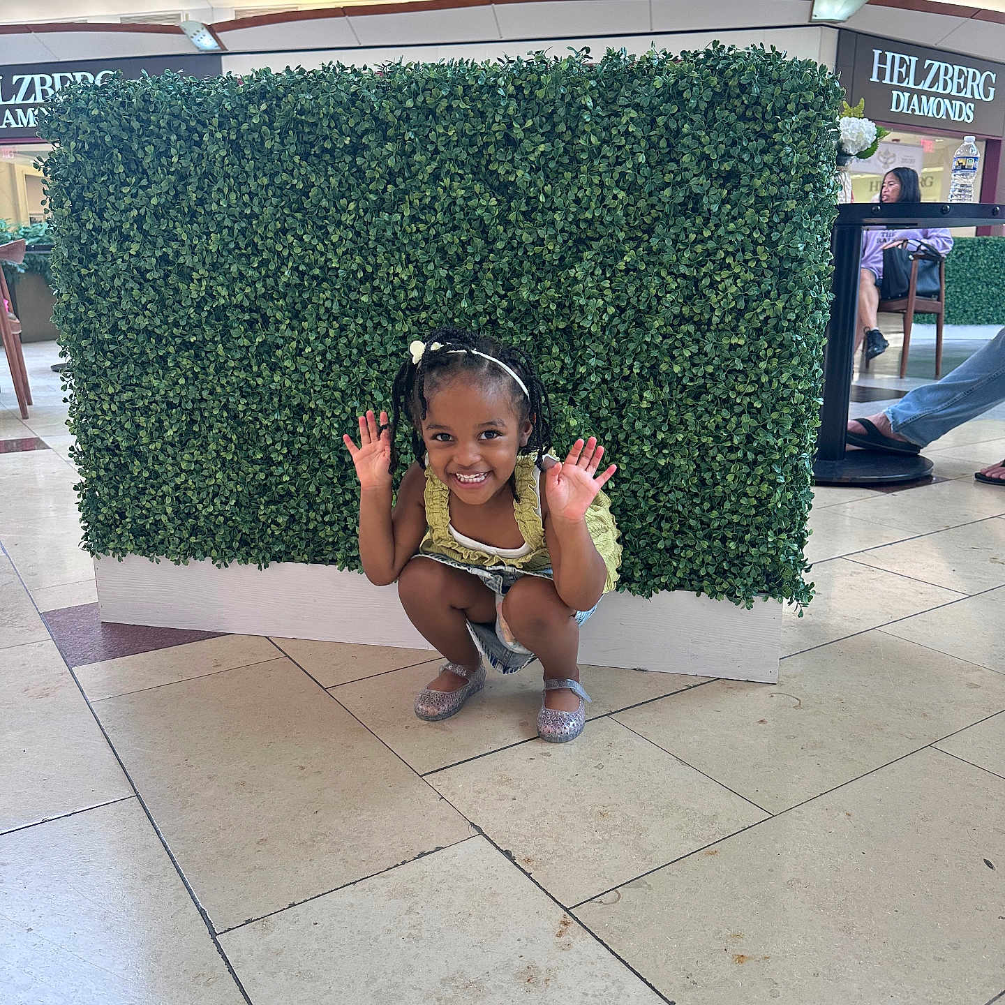 Zariah is registered to the contest to win money with this photo: background, casual_clothing, chair, child, floor_tiles, footwear, green_hedge, happy, headband, indoor, mall, person, playful, retail_store, seated, shoes, smiling, squatting, table, woman