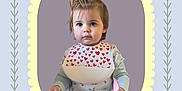 Valéria participe au concours pour gagner de l'argent avec cette photo : toddler, child, bib, plate, spoon, hair, pony_tail, heart_pattern, long_sleeve_shirt, cherry_pattern, portrait, indoor, cute, young_child, face, expression, holding, frame, decorative_border, pastel_colors