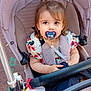 child, toddler, pacifier, stroller, plush_toy, baby, outdoor, cute, colorful_clothing, young_child, seatbelt, portrait, person, baby_carriage, sitting, curly_hair, playful, daylight, closeup, baby_fashion