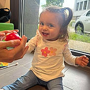 Eva a rejoint le concours — aidez-le/la à gagner de superbes lots ! casual_clothing, child, cute, daylight, floor, hand, happy, home, indoor, interaction, jeans, long_sleeve_shirt, person, playful, ponytail, red_toy, smile, toddler, toy, window