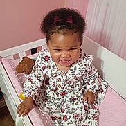 Mich'A is registered to the contest to win money with this photo: baby, child, smiling, portrait, crib, nursery, pink, floral_dress, dress, hair_bow, curly_hair, teddy_bear, toy, sitting, indoor, bedroom, blanket, yellow_socks, happy, photography