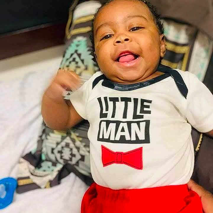 Deontavis is registered to the contest to win money with this photo: baby, child, smiling, red_pants, white_shirt, text_on_clothing, indoor, cushion, patterned_blanket, portrait, happy, infant, person, cute, tongue_out, clothing, relaxed, lying_down, home, face
