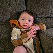 Eddie is registered to the contest to win money with this photo: baby, infant, child, plaid_clothing, hooded_outfit, couch, gray_couch, indoor, hands, face, blue_eyes, dark_hair, seated, cozy, cute, portrait, skin, expression, person, casual