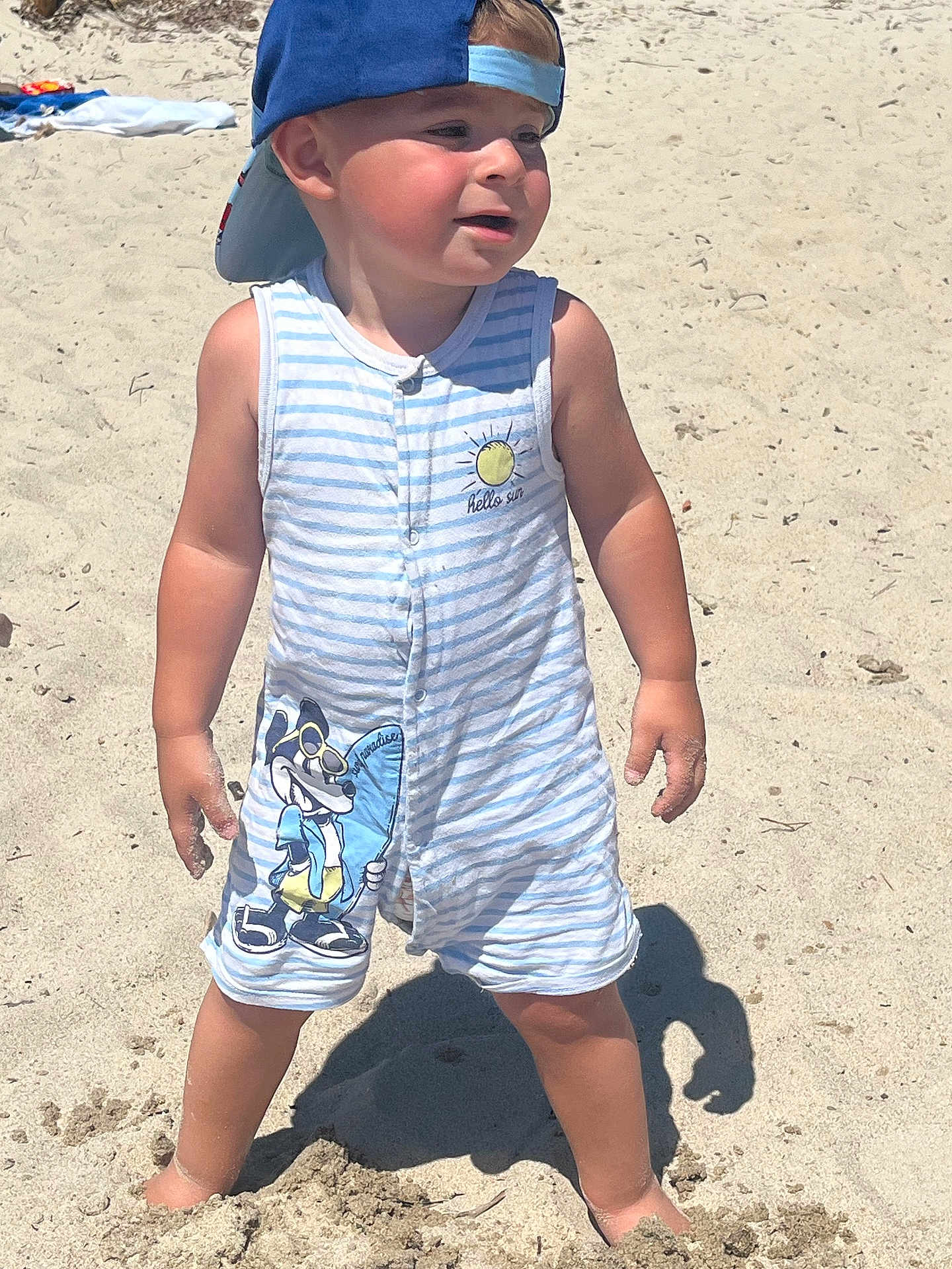 Bastien a rejoint le concours — aidez-le/la à gagner de superbes lots ! toddler, child, beach, sand, sunny, cap, romper, striped_clothing, cartoon_character, barefoot, summer, outdoor, young_child, cute, sunlight, shadow, playful, vacation, fun, sand_on_hands