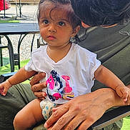 Mahi is registered to the contest to win money with this photo: toddler, child, adult, outdoor, sky, string_lights, chair, hand, face, curious, clothing, short_sleeve, hair, greenery, table, umbrella, foot, diaper, person, sunlight