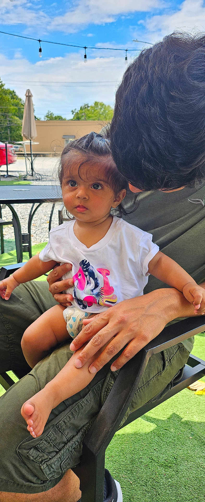 Mahi is registered to the contest to win money with this photo: toddler, child, adult, outdoor, sky, string_lights, chair, hand, face, curious, clothing, short_sleeve, hair, greenery, table, umbrella, foot, diaper, person, sunlight