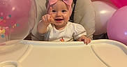 Kensli is registered to the contest to win money with this photo: baby, child, high_chair, pink_bow, birthday, balloon, party, celebration, smiling, pointing, indoor, happy, face, headwear, festive, toddler, clothing, table, person, decorations