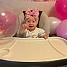 baby, child, high_chair, pink_bow, birthday, balloon, party, celebration, smiling, pointing, indoor, happy, face, headwear, festive, toddler, clothing, table, person, decorations