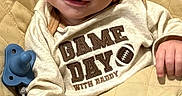 Jackson is registered to the contest to win money with this photo: baby, child, infant, smiling, portrait, indoors, seated, quilted_surface, blanket, pacifier, football, sportswear, clothing, hands, face, brown_hair, happy, cozy, close_up, photography