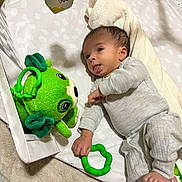 Jackson is registered to the contest to win money with this photo: baby, infant, child, lying_down, tongue_out, funny_expression, portrait, face, smiling, toy, frog_toy, green_toy, rattle, play_mat, blanket, bedroom, indoor, clothing, gray_outfit, cozy