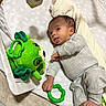 baby, infant, child, lying_down, tongue_out, funny_expression, portrait, face, smiling, toy, frog_toy, green_toy, rattle, play_mat, blanket, bedroom, indoor, clothing, gray_outfit, cozy